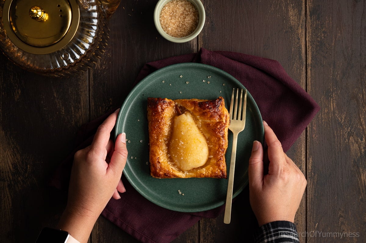 Someone holding a green plate with a baked spiced pear in golden brown puff pastry sprinkled with coarse sugar.