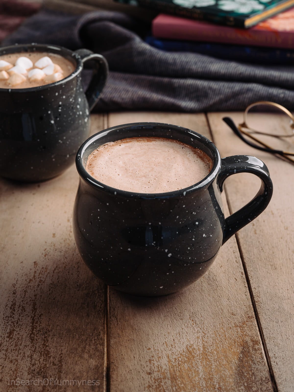 A black speckled mug full of hot chocolate on a light wooden table next to another mug and a pair of glasses.