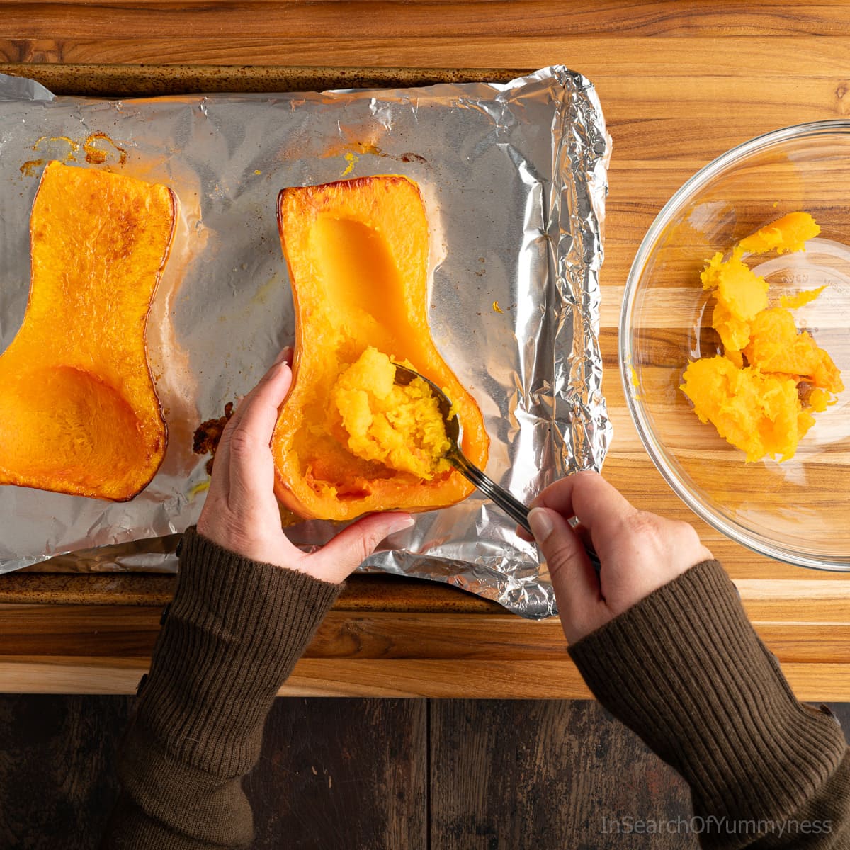 Someone using a spoon to scoop out roasted butternut squash from its skin. There's cooked squash in a clear bowl as well.