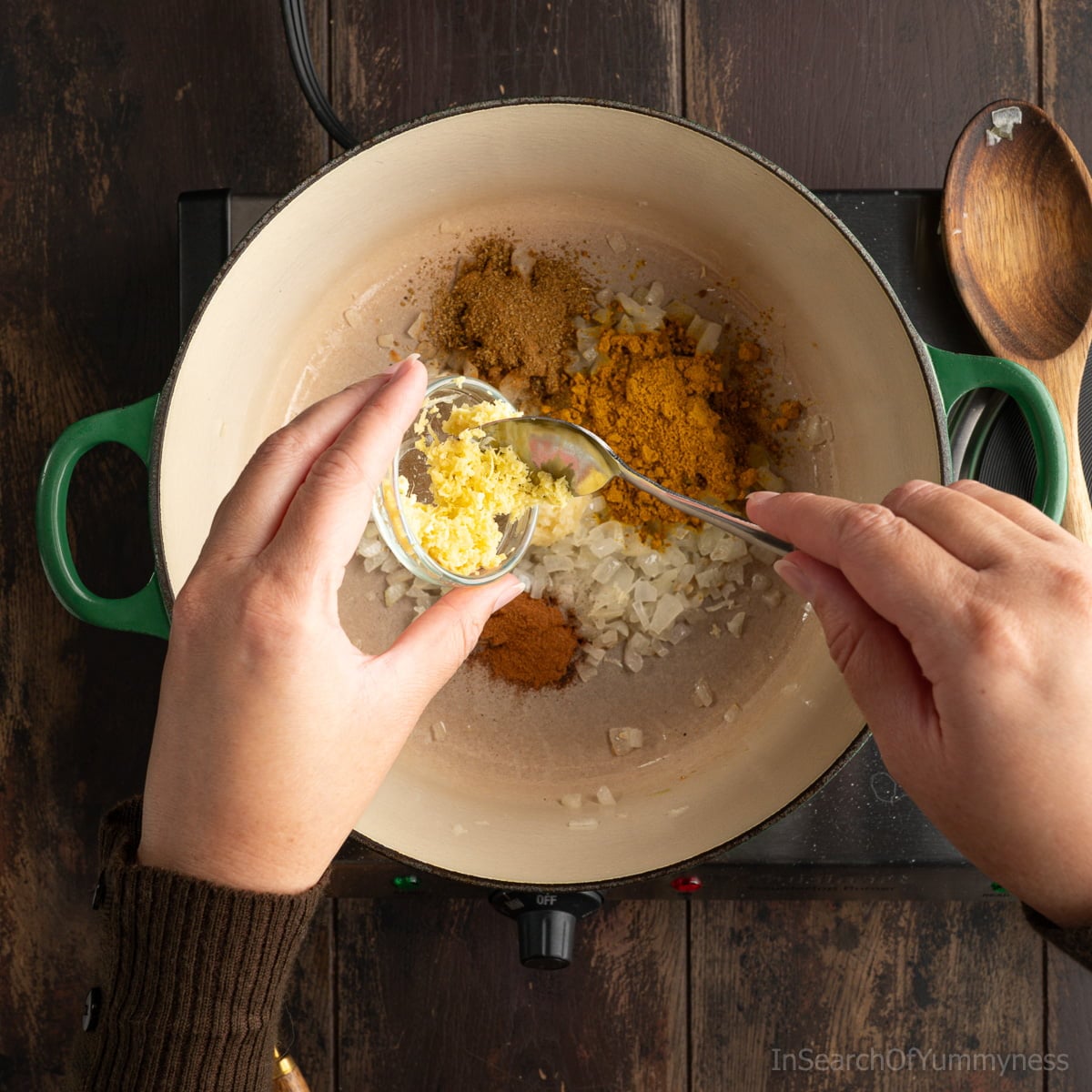 Someone adding grated ginger from a small glass bowl into a green Le Creuset Dutch oven that already has spices added to the onions in it.