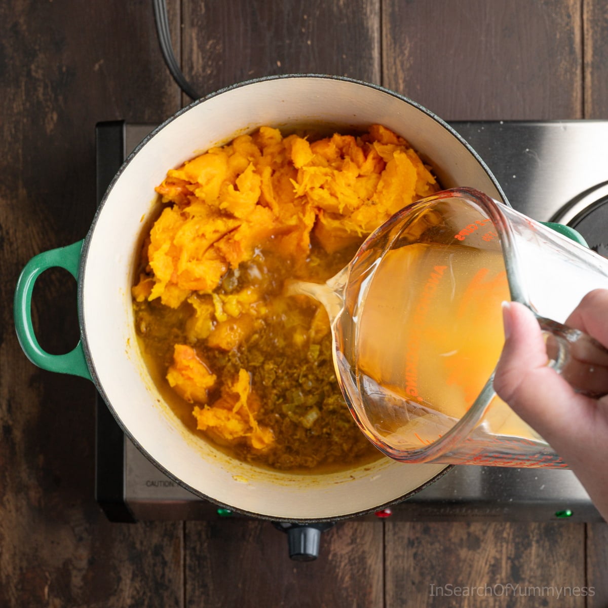 Someone adding vegetable stock from a Pyrex measuring jug to a green Le Creuset Dutch oven full of cooked squash, onions and spices.