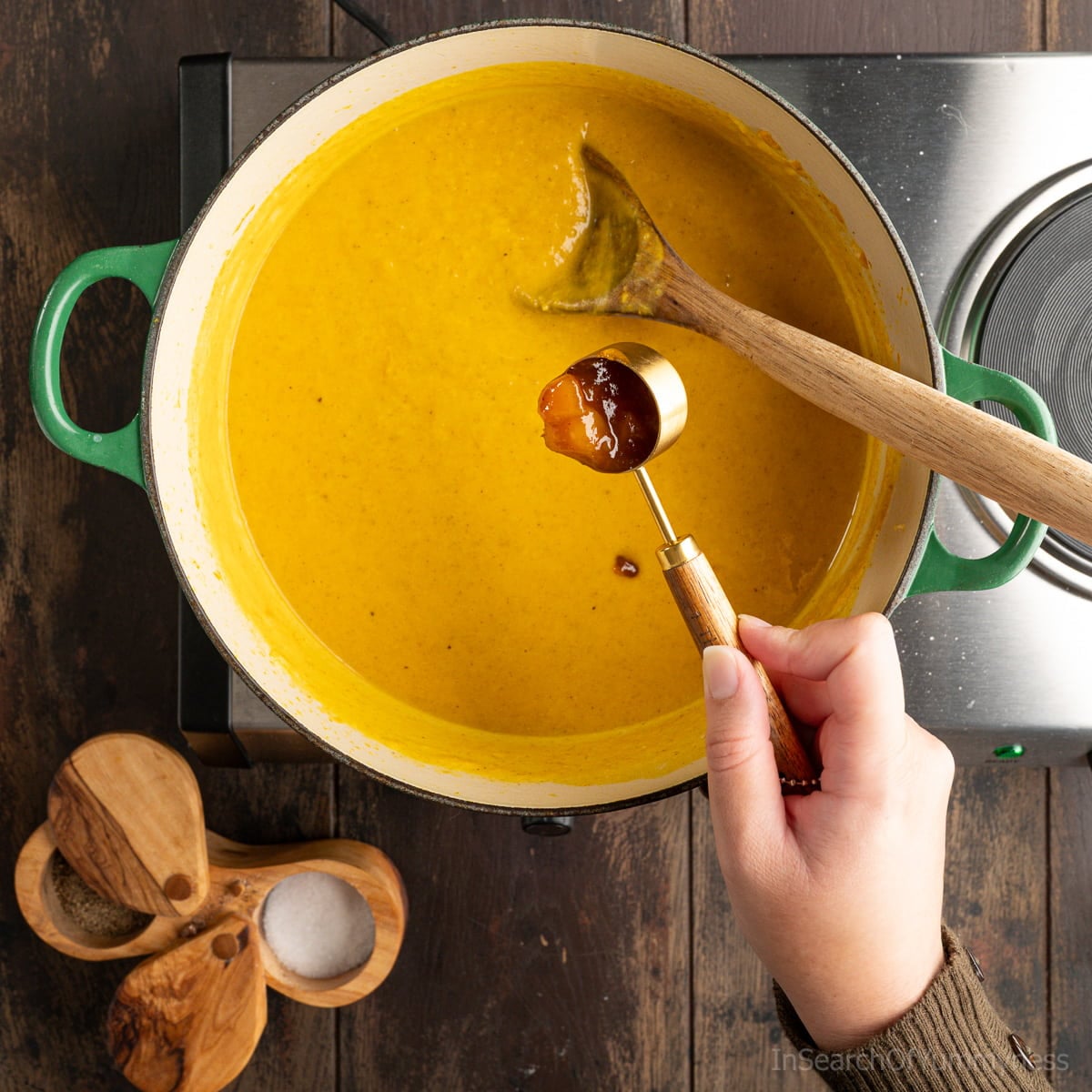 Someone adding a tablespoon of mango chutney to a green Dutch oven filled with curried butternut squash soup.