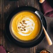 An overhead view of a vibrant yellow bowl of butternut squash soup garnished with pumpkin seeds and a swirl of coconut milk.