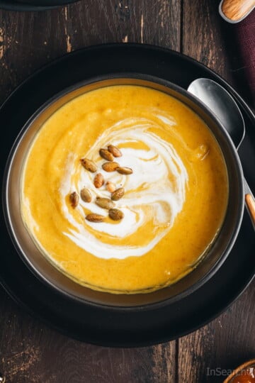 An overhead view of a vibrant yellow bowl of butternut squash soup garnished with pumpkin seeds and a swirl of coconut milk.