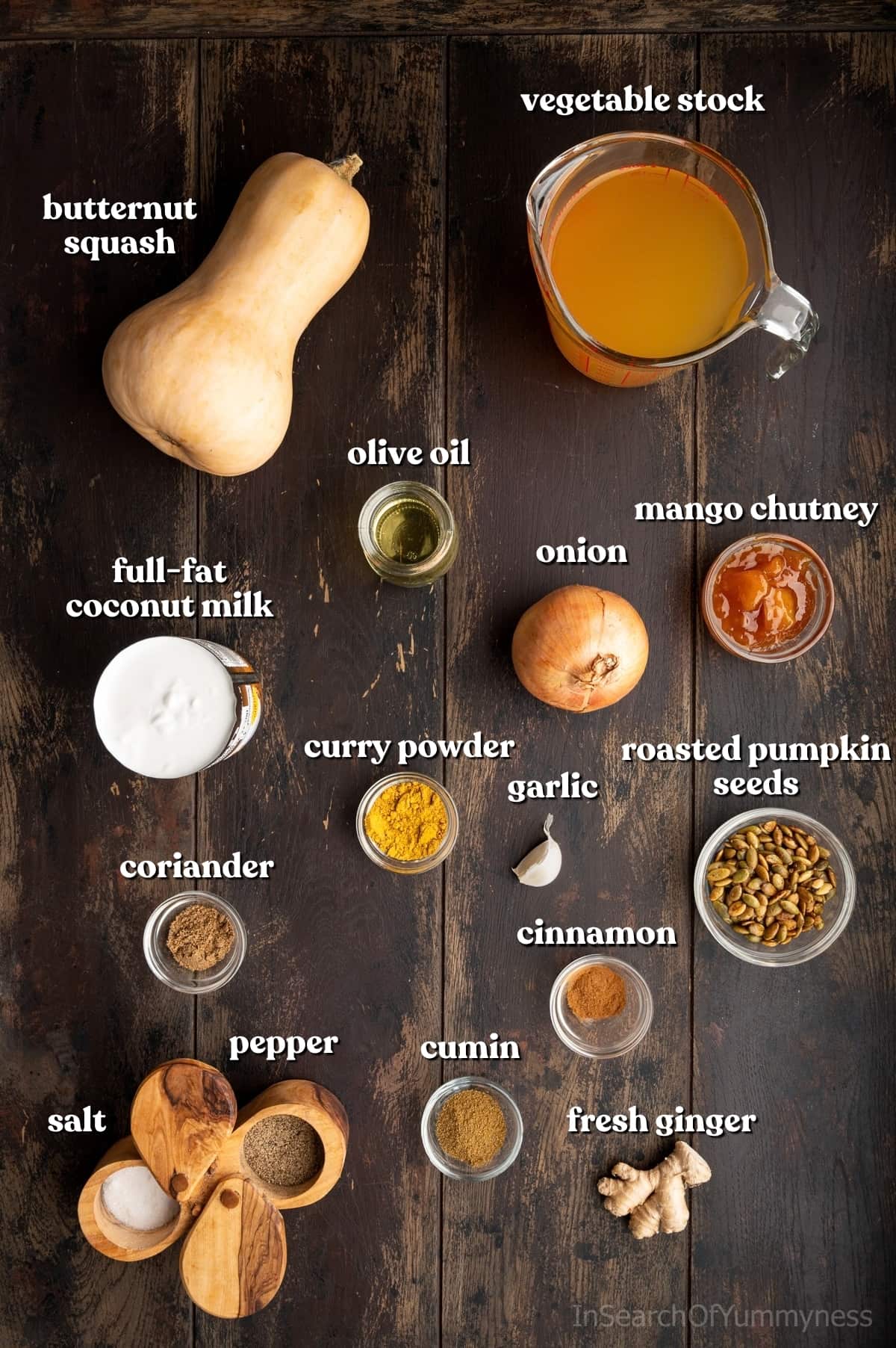 Ingredients for curried butternut squash soup, including butternut squash, vegetable stock, coconut milk, onion, garlic, ginger, spices, and mango chutney on a dark wooden table.