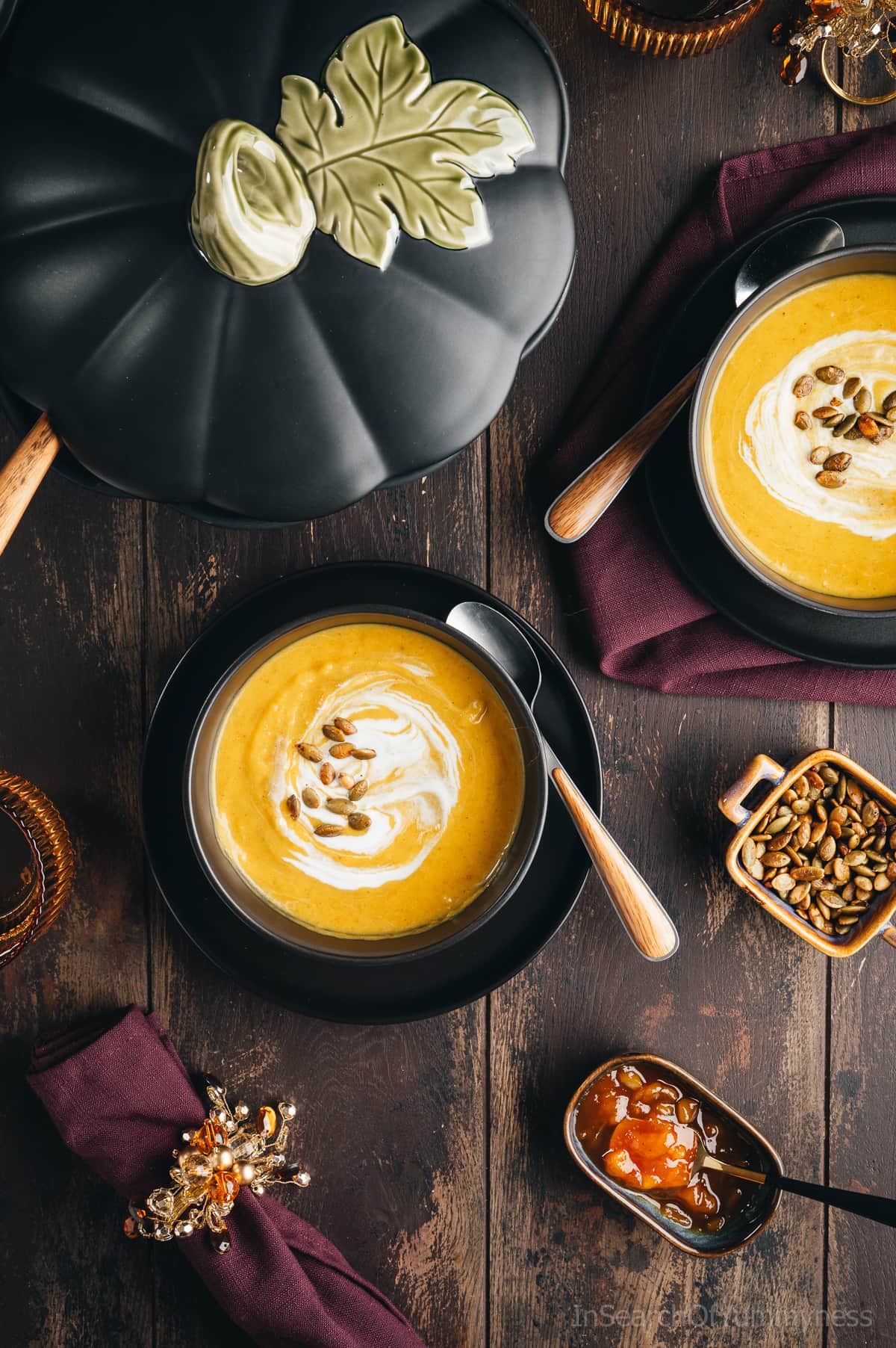 Two bowls of vibrant yellow curried butternut squash soup in black bowls, on black plates, next to a large pumpkin-shaped soup tureen. There are small pinch bowls with mango chutney and roasted pumpkin seeds next to the soup bowls, as well as purple napkins and ornate napkin rings.