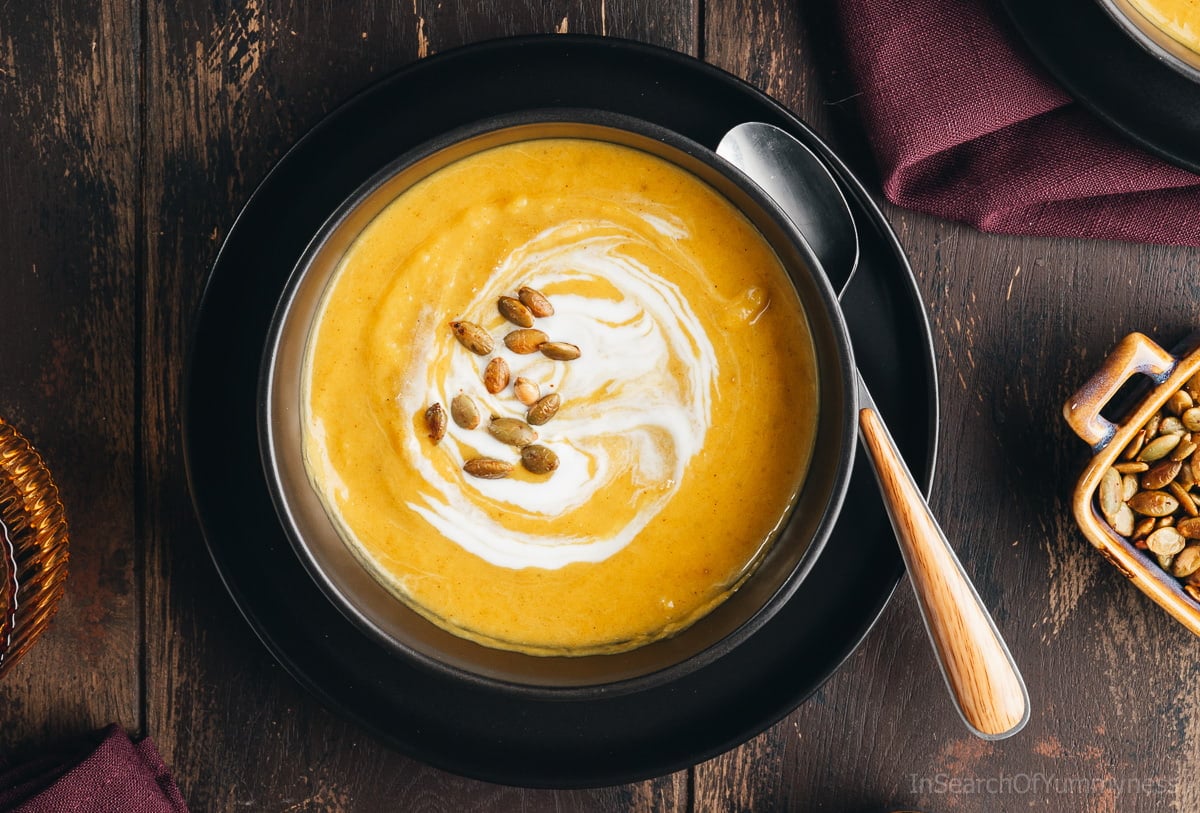 An overhead view of a vibrant yellow bowl of butternut squash soup garnished with pumpkin seeds and a swirl of coconut milk.