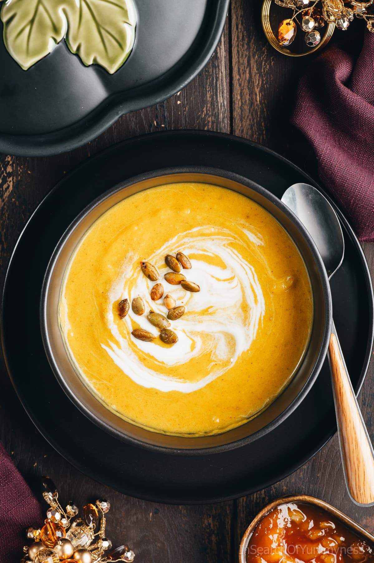 An overhead view of a vibrant yellow bowl of butternut squash soup garnished with pumpkin seeds and a swirl of coconut milk. There is a pumpkin soup tureen, two purple napkins, two bejewelled napkin holders and a pinch bowl of mango chutney next to the soup.