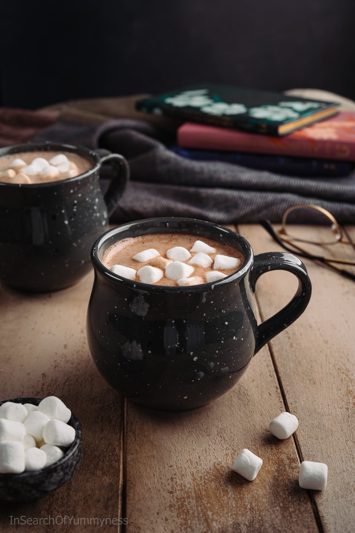 A black speckled mug full of hot chocolate, topped with mini marshmallows, on a light wooden table next to another mug and a pair of glasses.