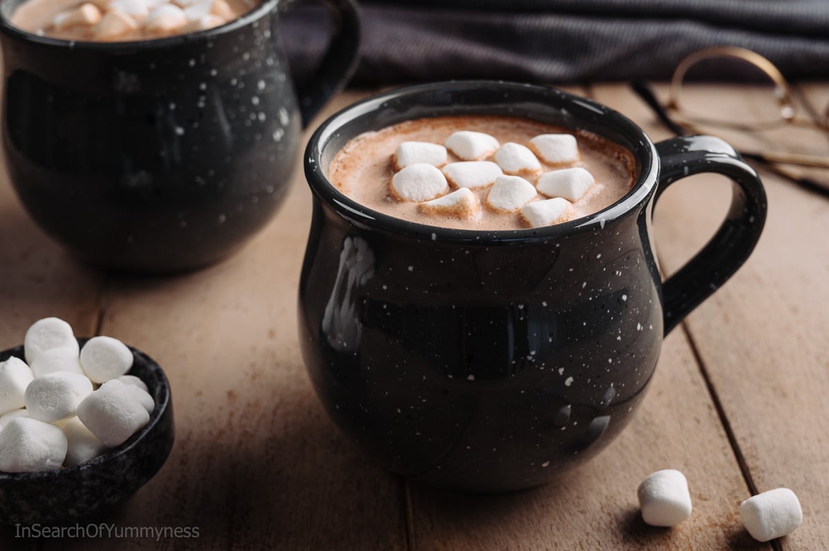 A black speckled mug full of hot chocolate, topped with mini marshmallows, on a light wooden table next to another mug and a pair of glasses.