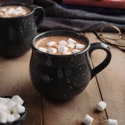 A black speckled mug full of homemade hot chocolate, topped with mini marshmallows, on a light wooden table next to a pair of glasses.