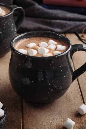 A black speckled mug full of homemade hot chocolate, topped with mini marshmallows, on a light wooden table next to a pair of glasses.