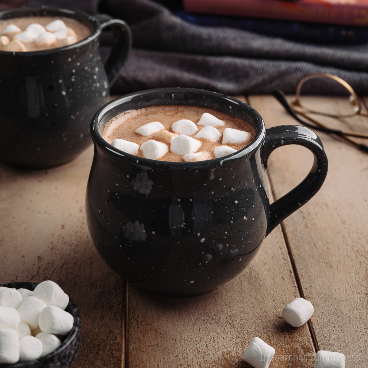 A black speckled mug full of homemade hot chocolate, topped with mini marshmallows, on a light wooden table next to a pair of glasses.