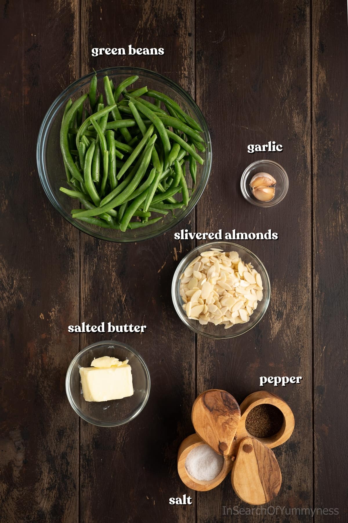 Ingredients for green beans almondine, including green beans, garlic, slivered almonds, salted butter, salt and pepper, on a dark wooden table.
