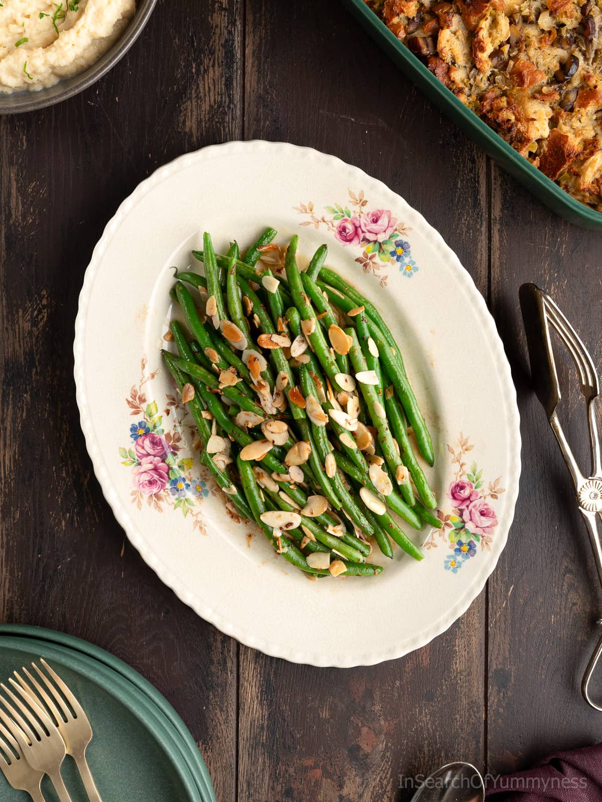A vintage floral plate with green beans almondine; buttery sautéed green beans topped with toasted slivered almonds. There are other holiday side dishes around the beans, plus a stack of green plate, gold forks, and silver serving tongs.