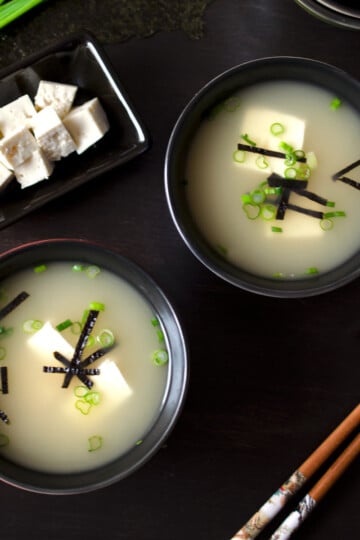 Two small bowls of miso soup made without dashi on a dark brown table next to a black plate with diced tofu and a pair of chopsticks.