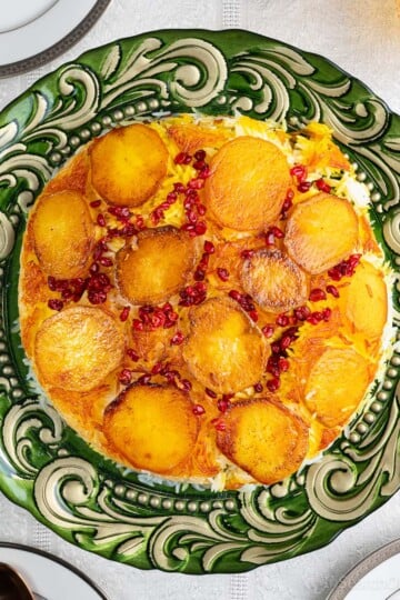 An ornate green and gold platter with potato tahdig: crispy golden brown potatoes over buttery saffron-stained basmati rice, topped with bright red barberries. There are spoons, forks and white plates around the platter, and a gold-tinted water glass.