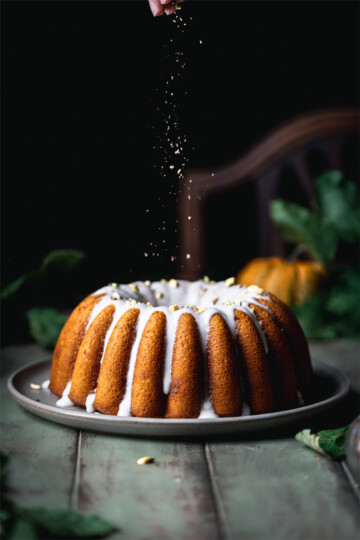 Sprinkling nuts over a beautiful pumpkin bundt cake on a table surrounded by greenery.