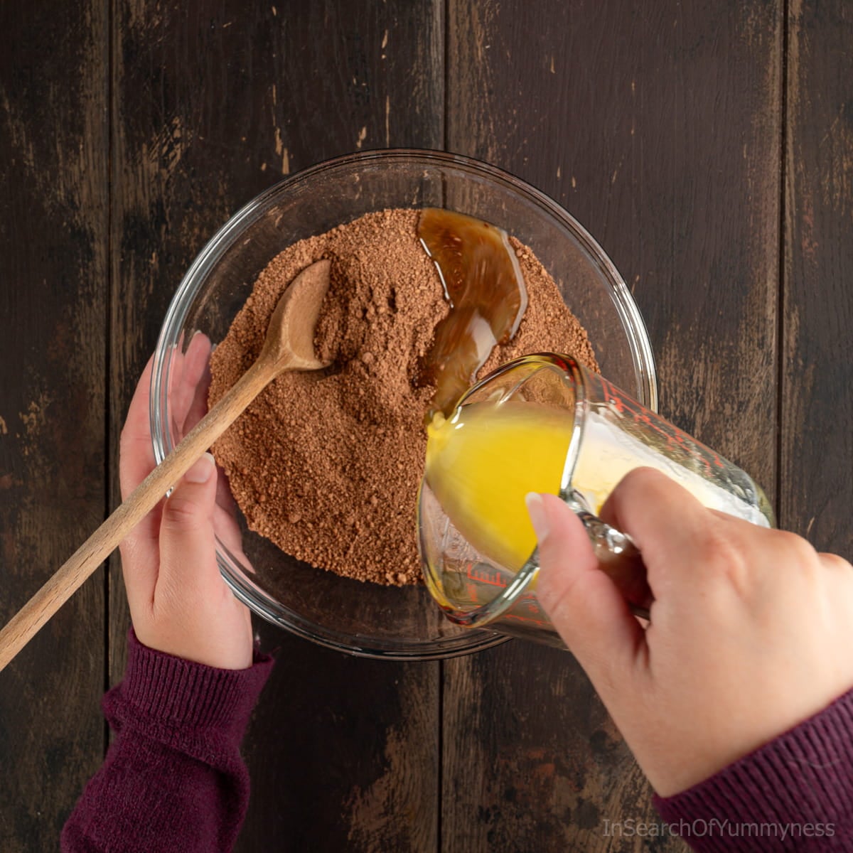 Pouring melted butter from a small Pyrex measuring jug into a clear bowl with chocolate graham crumbs. There's a wooden spoon in the bowl.