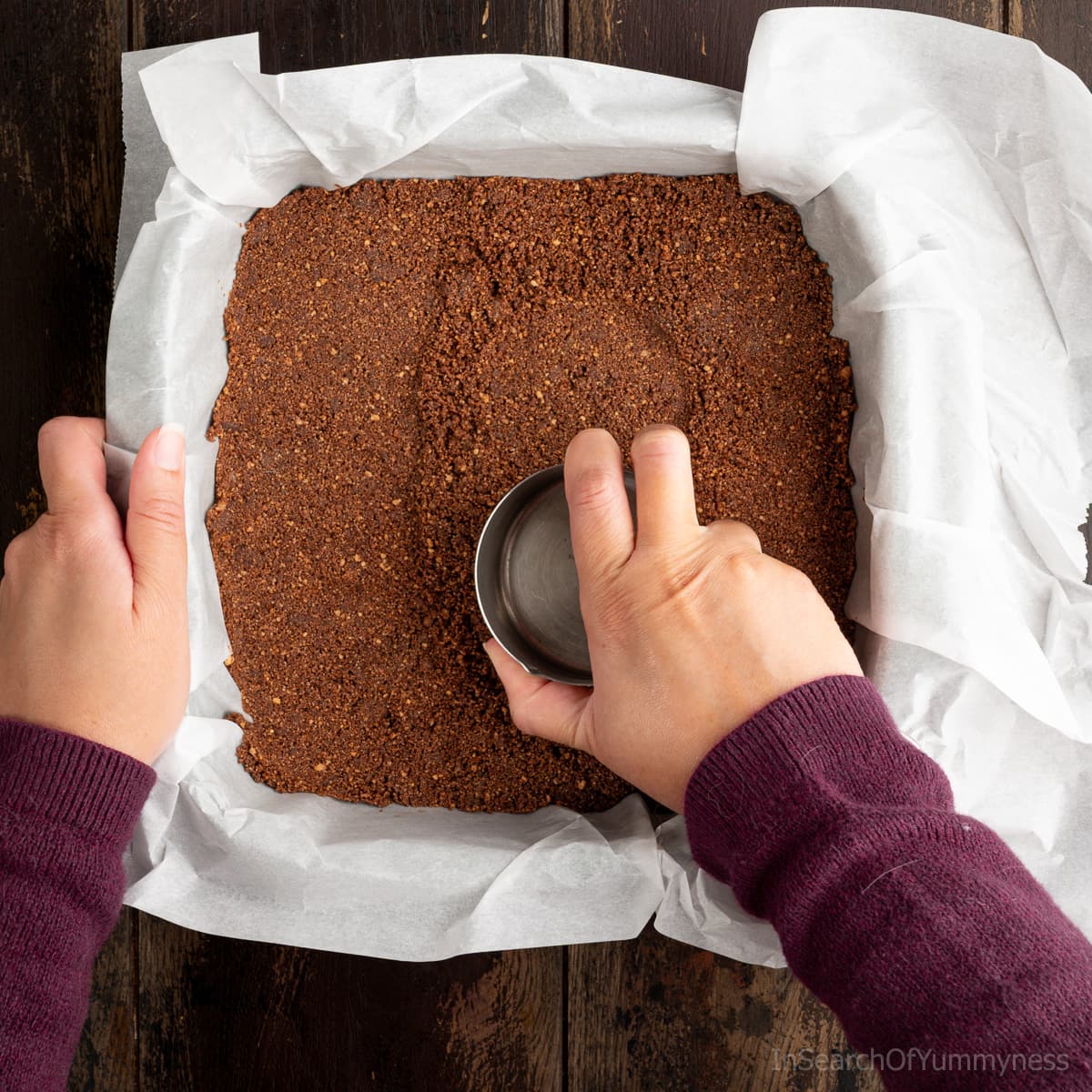 Using the back of a stainless steel mesuring cup to press down chocolate graham crumbs mixed with butter to make the cheesecake bar crust.