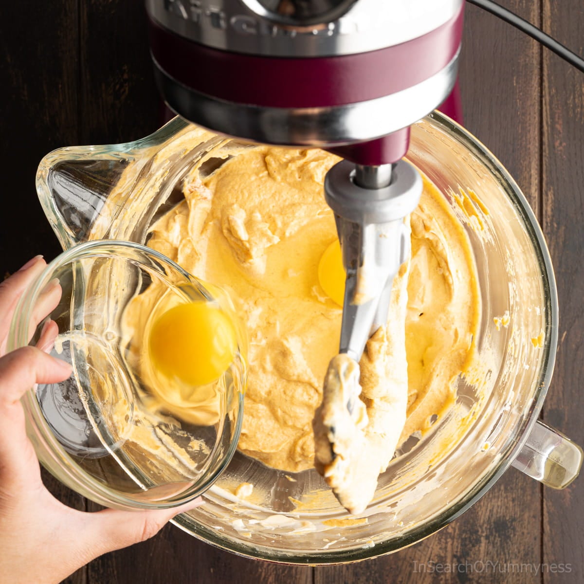 Adding two cracked eggs to the pumpkin cheesecake filling in the mixing bowl of a KitchenAid stand mixer.