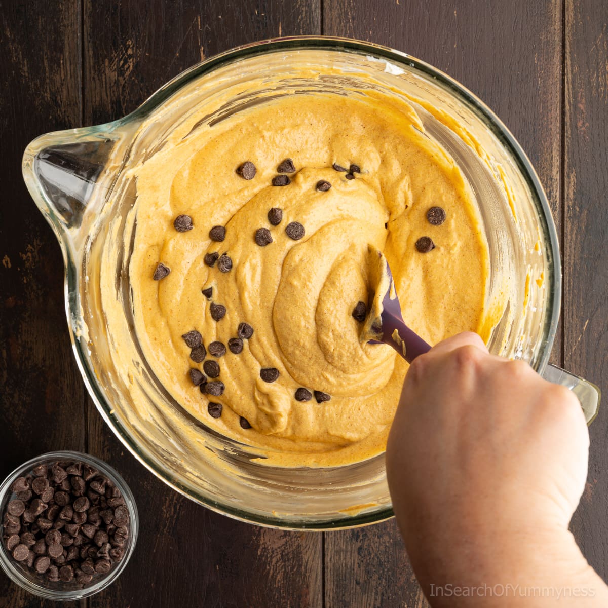 Using a spatula to stir semi-sweet chocolate chips into the mixed pumpkin cheesecake filling in a clear stand mixer bowl.