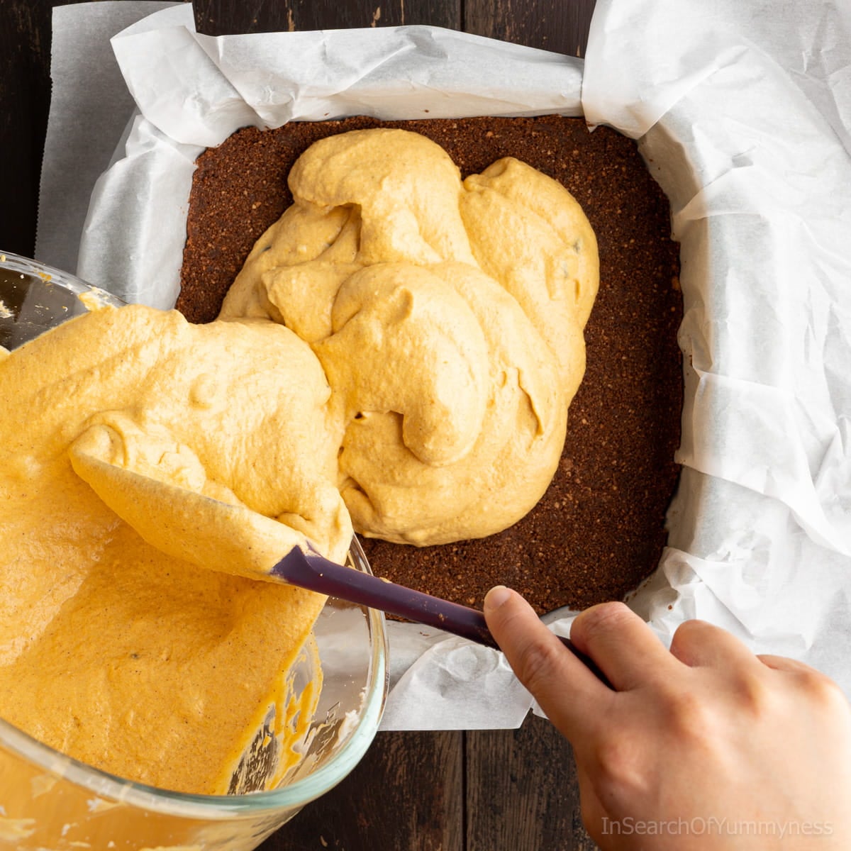 Scraping the mixed pumpkin cheesecake filling from the mixer bowl into the baking dish with the chocolate graham crumb crust. The baking dish is lined with parchment paper.
