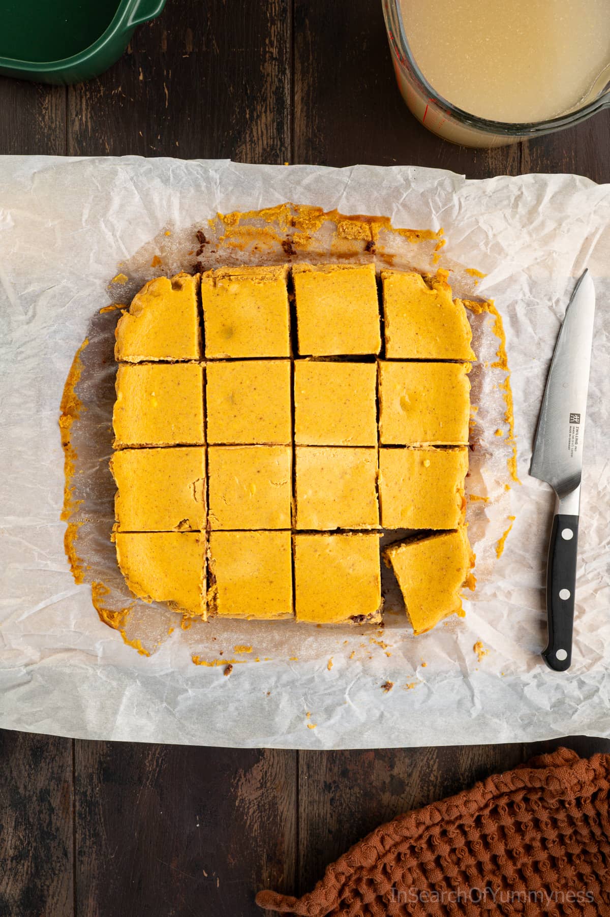 Sixteen sliced pumpkin cheesecake bars on crumpled parchment paper, next to a knife, a brown towel, and a Pyrex jug of water used to heat up the knife.