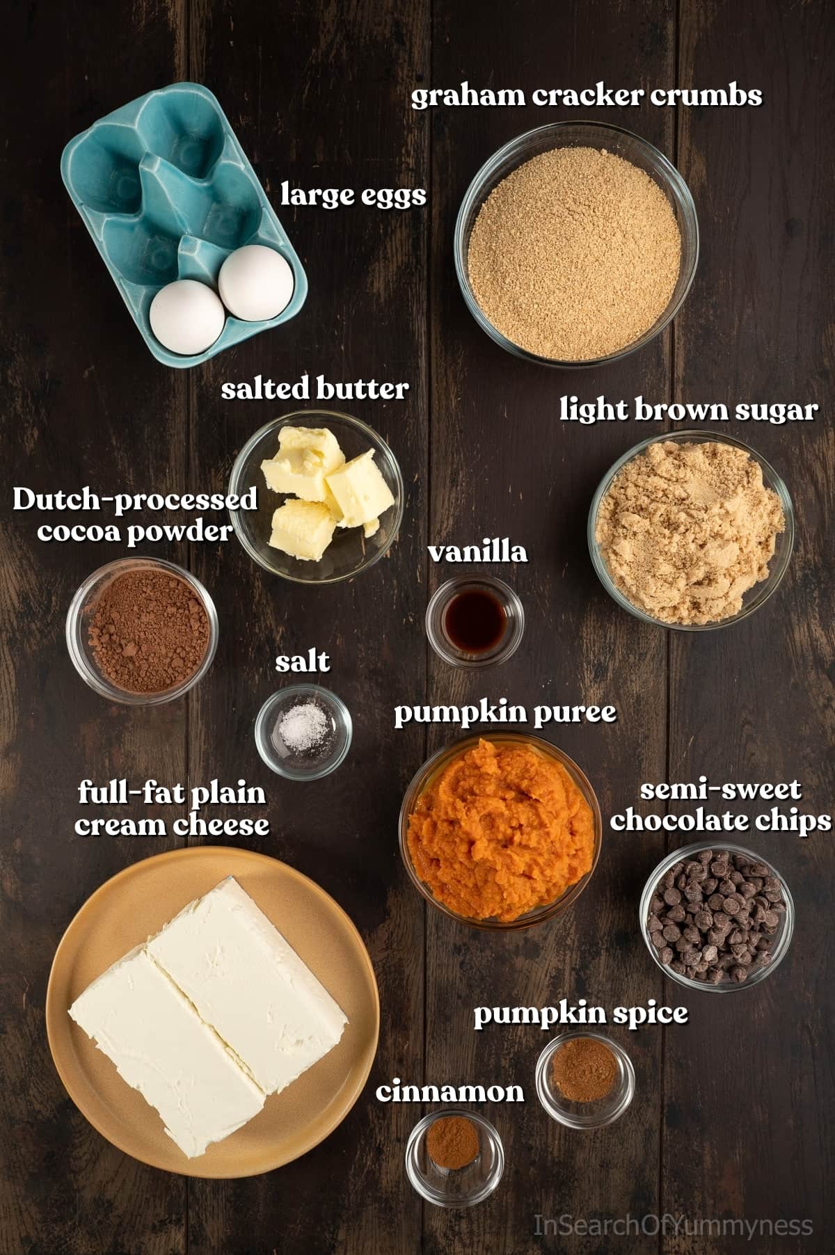 Ingredients for pumpkin cheesecake bars, including eggs, graham cracker crumbs, brown sugar, butter, pumpkin puree, cream cheese, pumpkin spice and more, on a dark brown table.