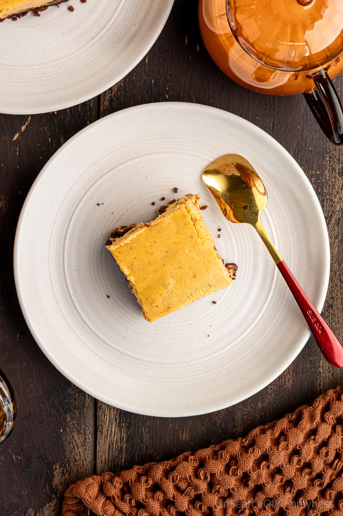 A pumpkin cheesecake bar studded with chocolate chips, with a dark brown chocolate graham crust, on a shallow white plate, on a dark brown table. There's a gold and red spoon and a pumpkin-shaped clear orange mug next to the plate.