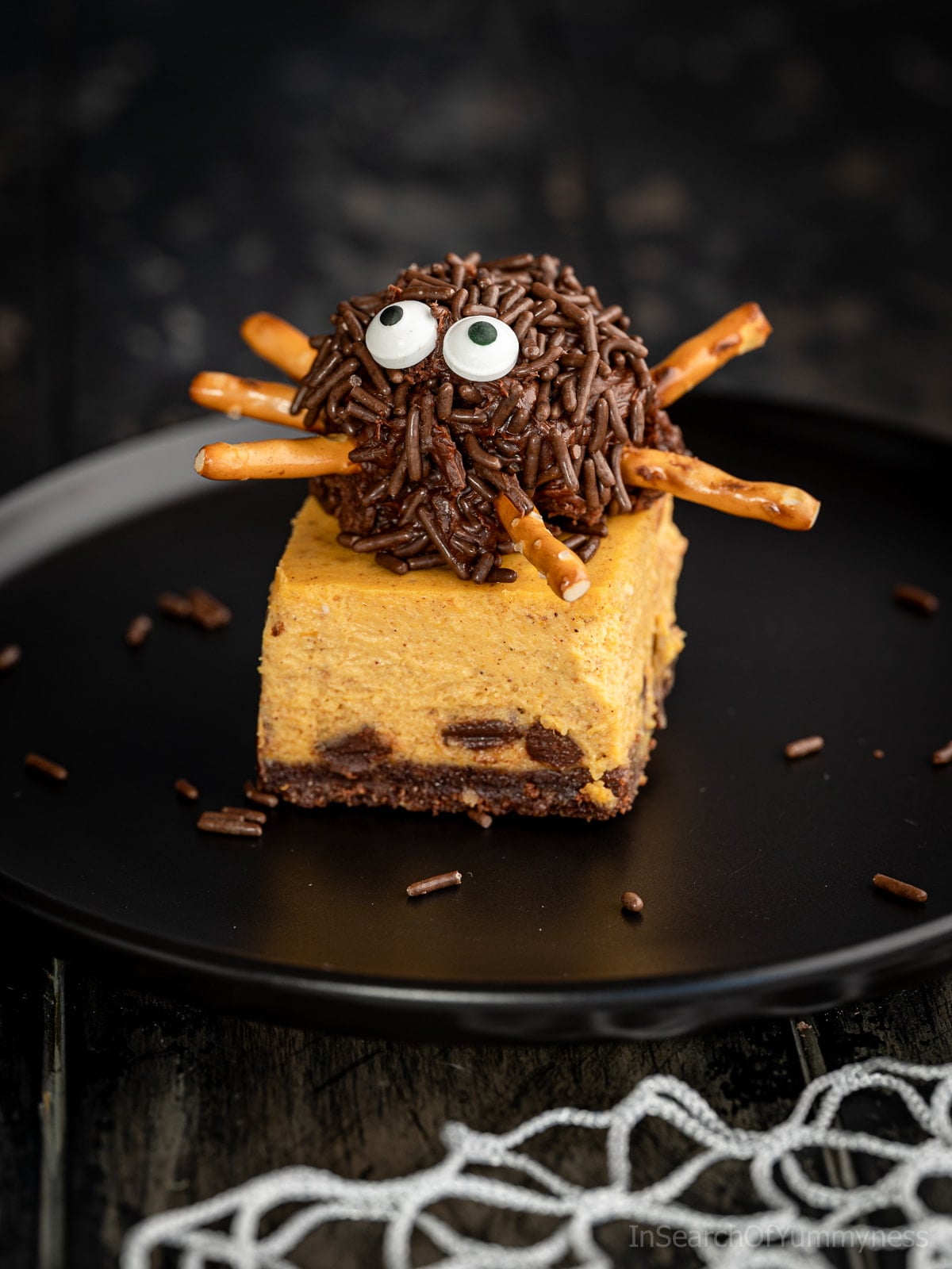 A cute spider brownie treat sitting on a pumpkin cheesecake bar on a black plate.