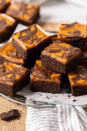 A pile of sliced pumpkin cheesecake brownies on a cooling rack with a white towel.