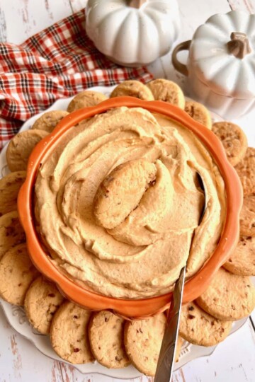 An orange pumpkin-shaped bowl full of pumpkin cream cheese dip, surrounded by cookies.