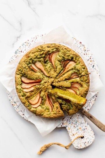 A whole pumpkin seed cake topped with pumpkin seeds and sliced red pears, with a slice taken out to show the bright green crumb.