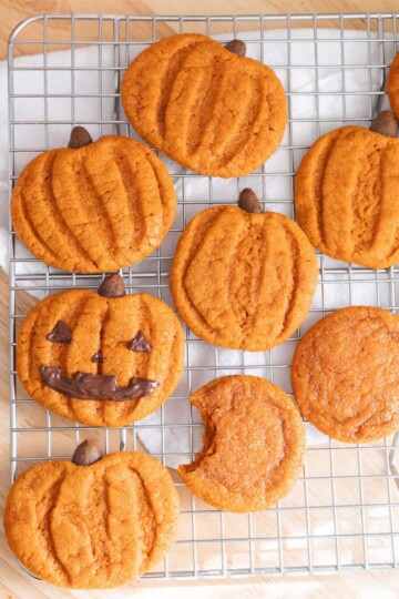 Cute pumpkin-shaped snickerdoodle cookies cooling on a wire rack. One is decorated like a jack-o'-lantern.