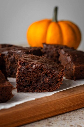 Moist pumpkin spice brownies on a piece of parchment on a cutting board, with a pumpkin behind it.
