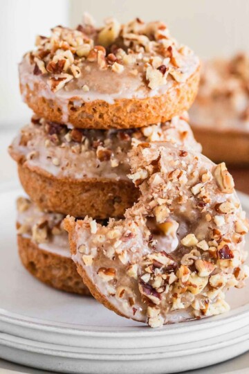 A pile of three pumpkin spice donuts topped with crushed nuts, with a fourth leaning on the side of the plate.